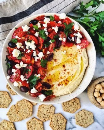 Bowl of greek salad hummus surrounded by crackers, parsley, and a small bowl of chickpeas