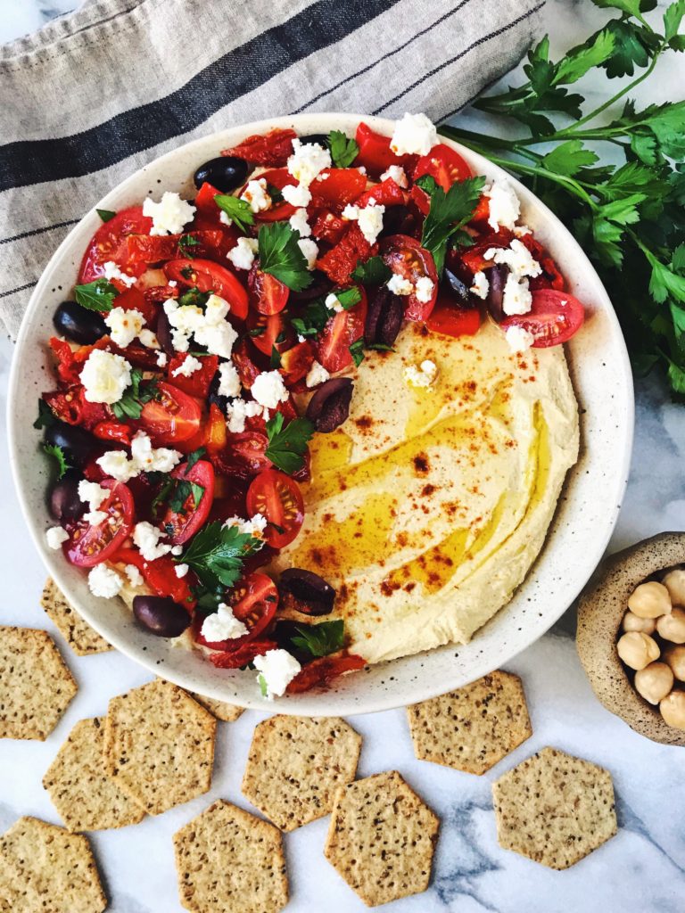 Bowl of greek salad hummus surrounded by crackers, parsley, and a small bowl of chickpeas