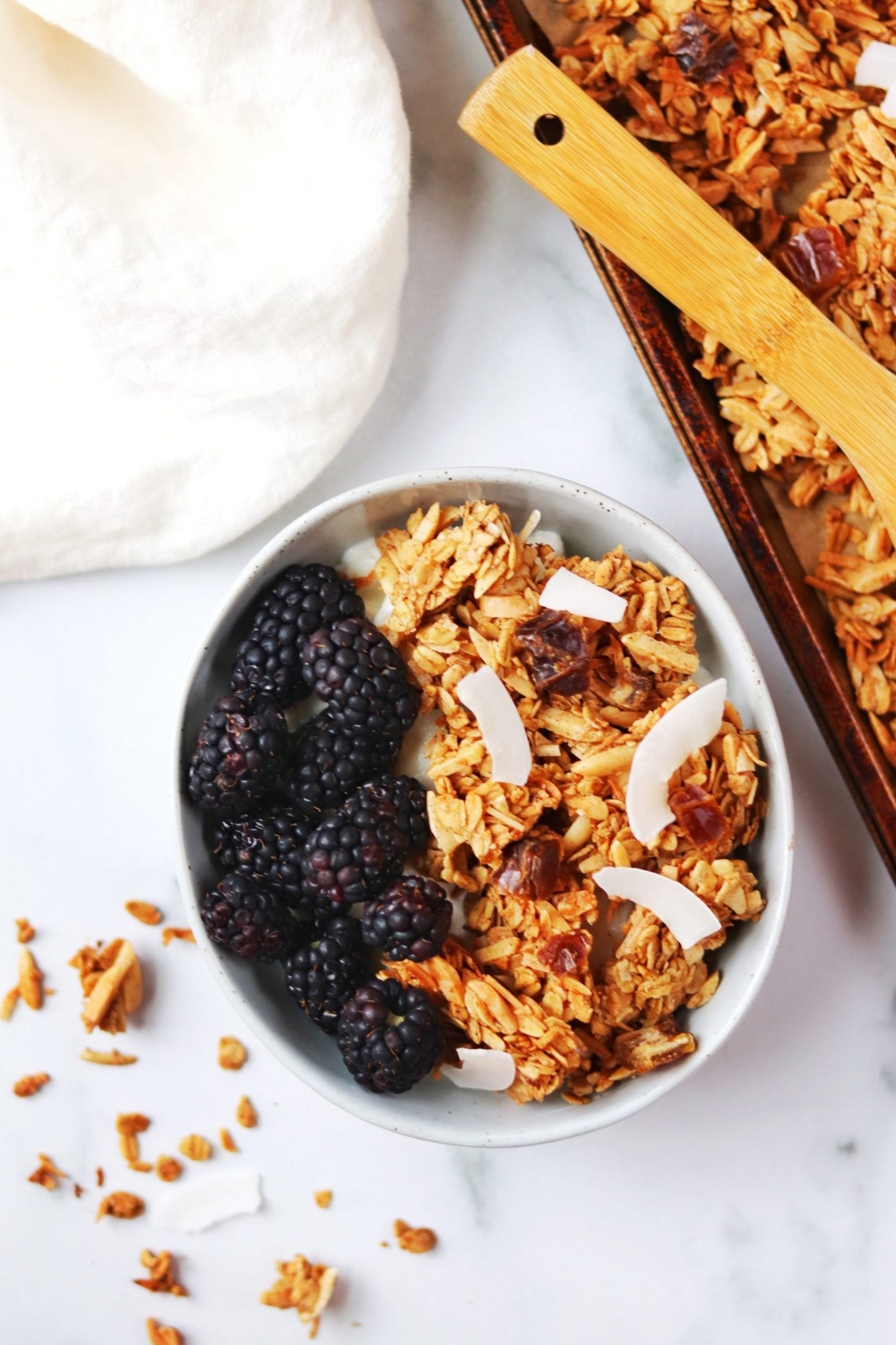 Bowl of granola with blackberries and coconut flakes, on a white backdrop with some granola scattered around