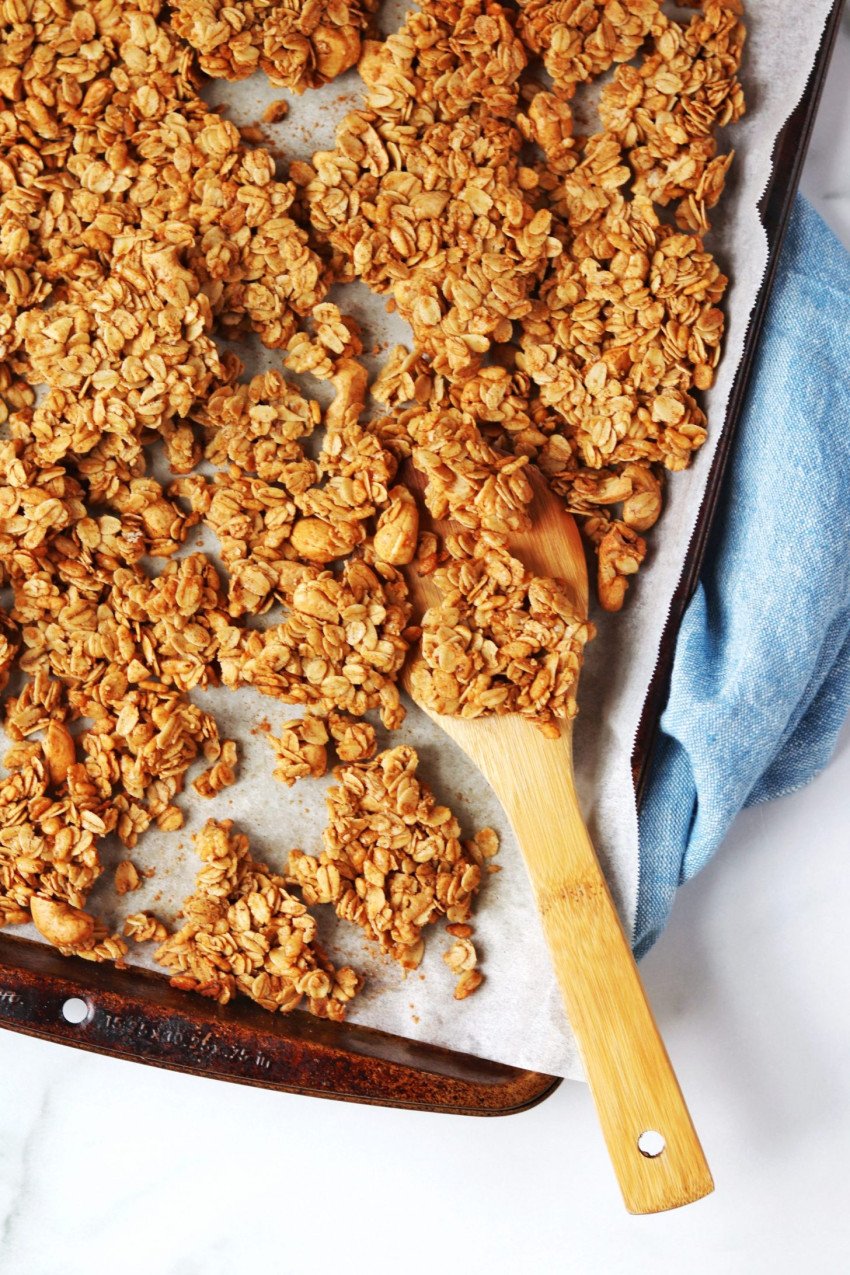 Baking tray with cinnamon granola, with a wooden mixing spoon and a blue dish towel