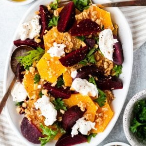 Citrus, beet and burrata salad on a white plate with gold serving spoons surrounded by bowls of ingredients and dressing