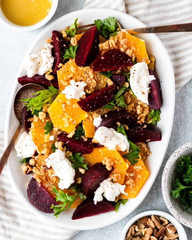 Citrus, beet and burrata salad on a white plate with gold serving spoons surrounded by bowls of ingredients and dressing