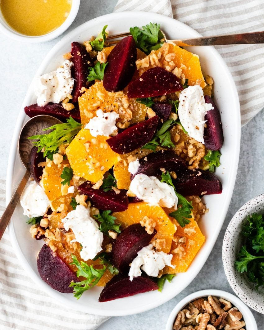 Citrus, beet and burrata salad on a white plate with gold serving spoons surrounded by bowls of ingredients and dressing