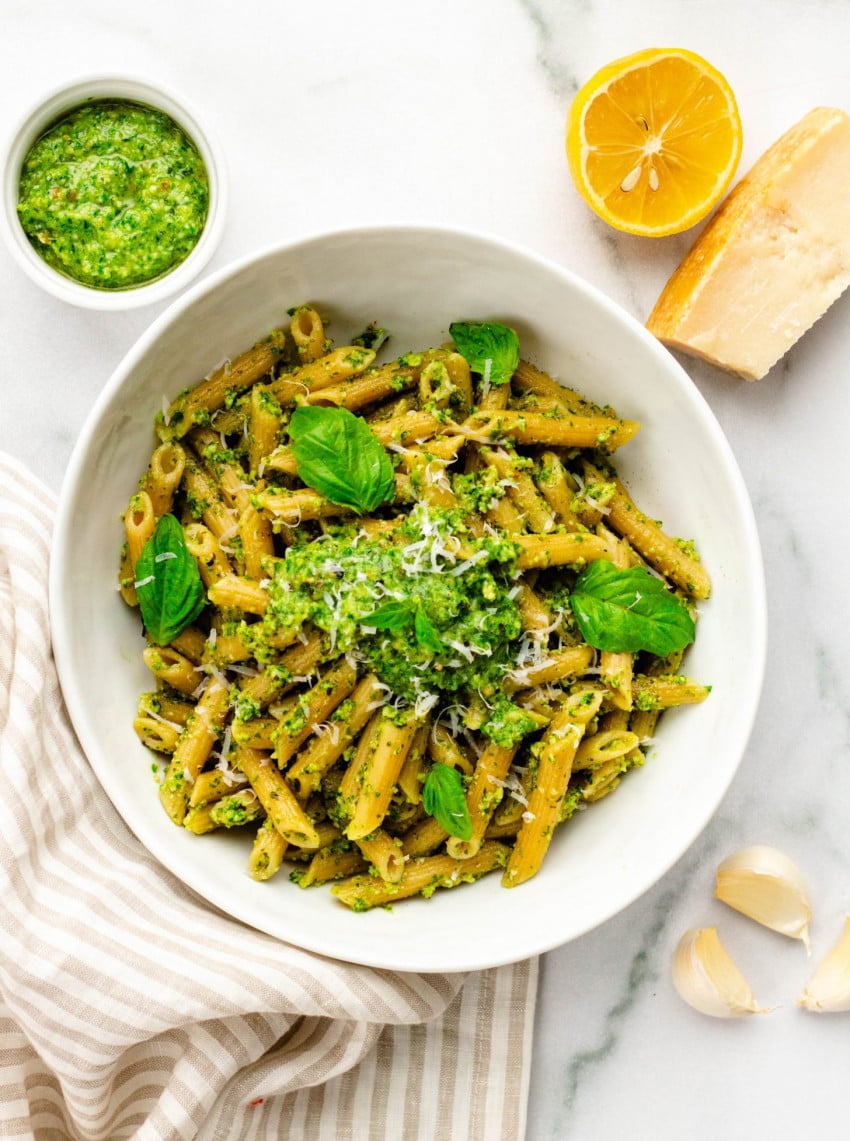 bowl of kale pesto pasta with ingredients surrounding it, on a white marble background
