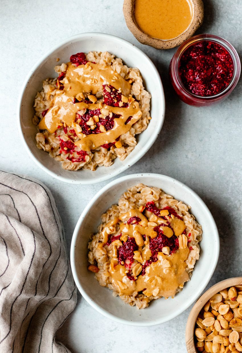 Overhead shot of two bowls of peanut butter and jelly oatmeal with small bowls of ingredients