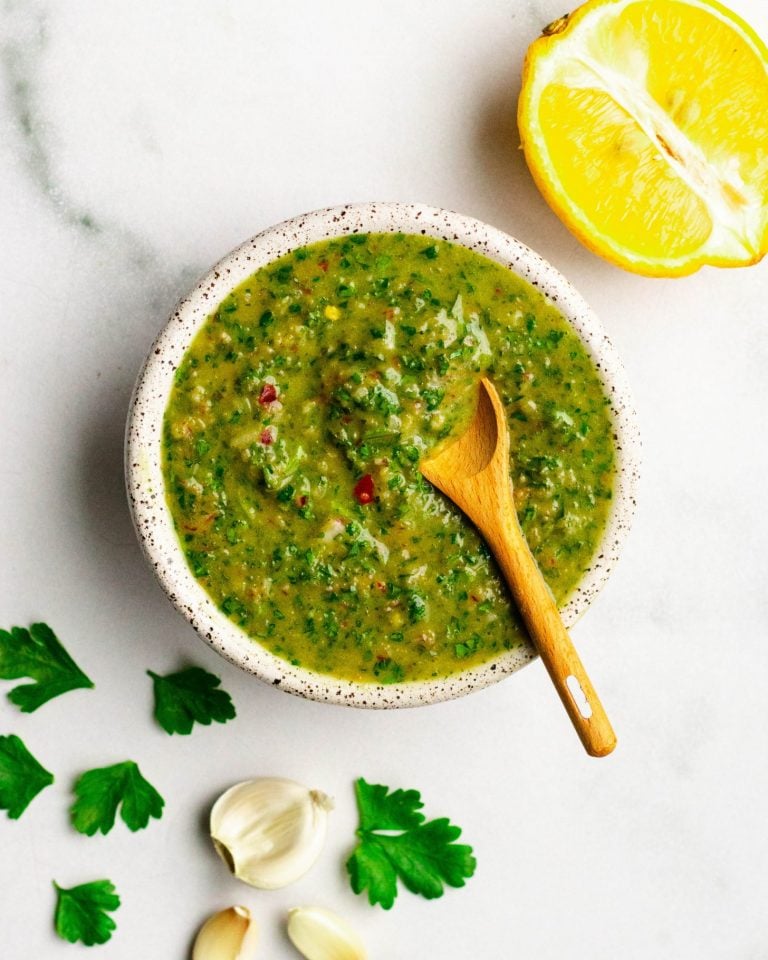 Homemade chimichurri sauce in a dish with small wooden spoon and ingredients surrounding