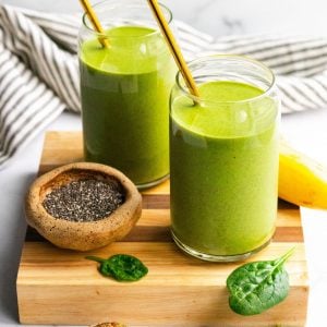two green smoothies in glasses with metal straws on a cutting board, ingredients around the smoothies