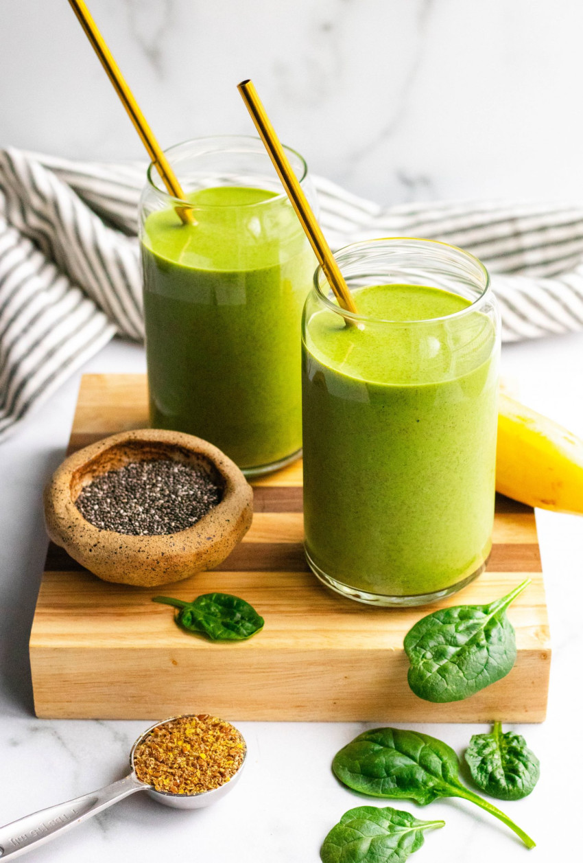 two green smoothies in glasses with metal straws on a cutting board, ingredients around the smoothies