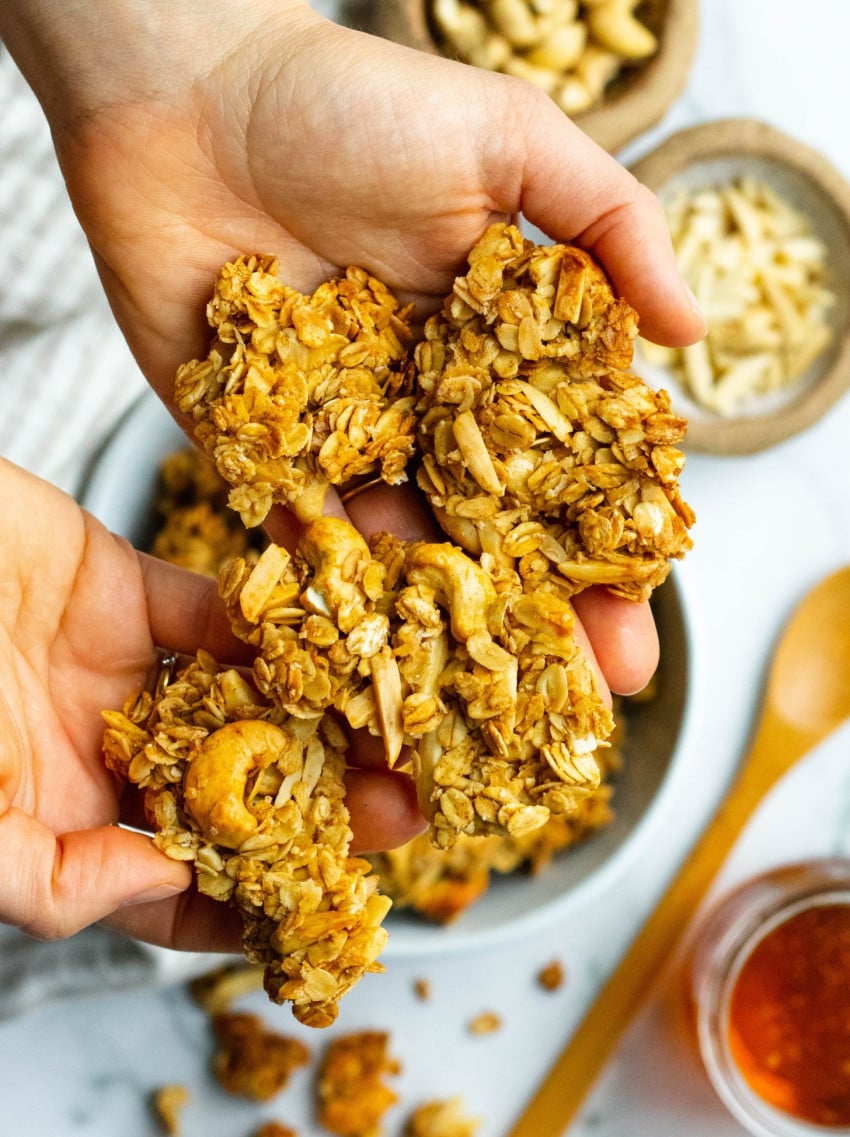Hands holding big clusters of honey nut granola with ingredients in the background