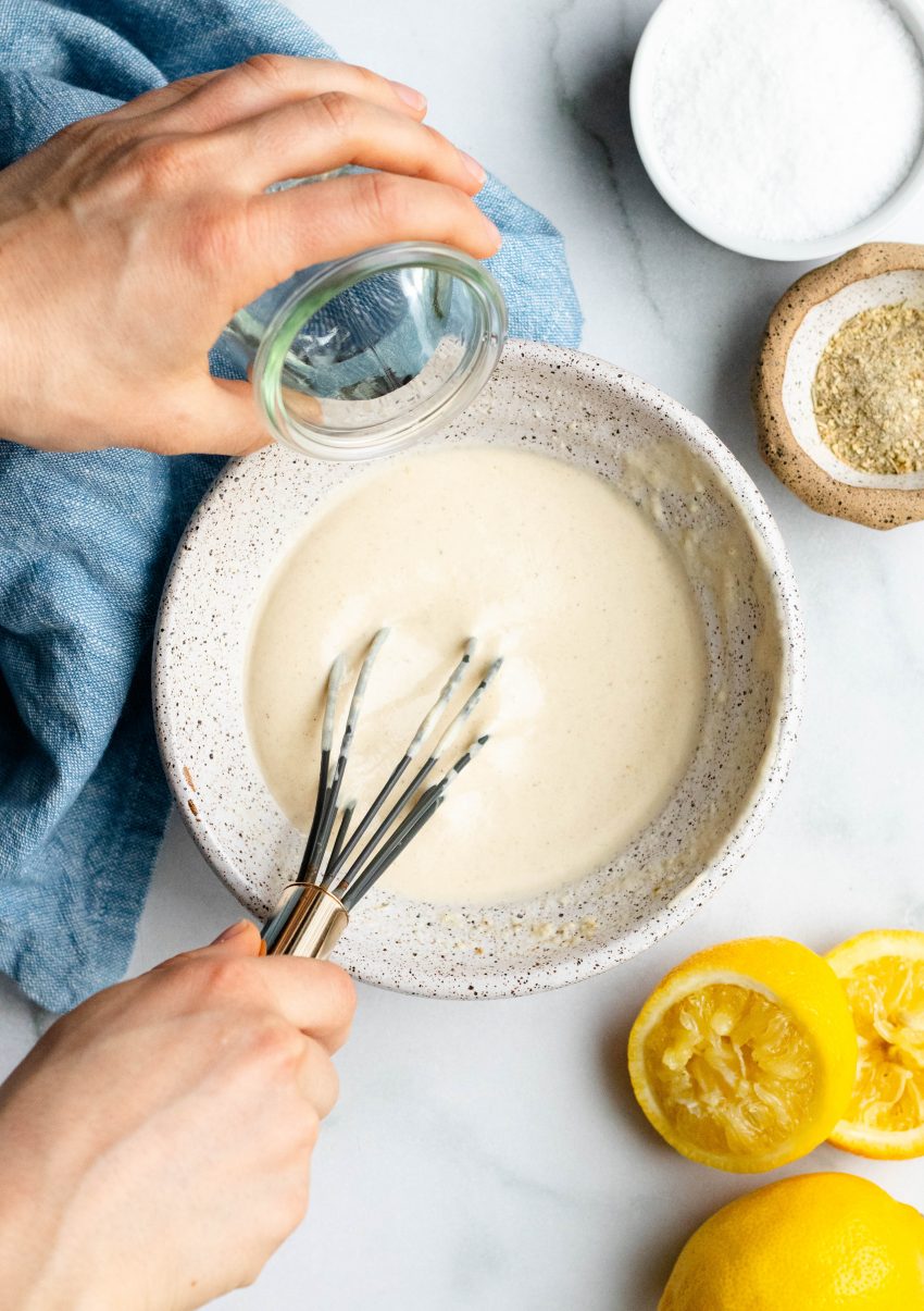 Hands adding water to and whisking lemon tahini sauce in a speckled bowl