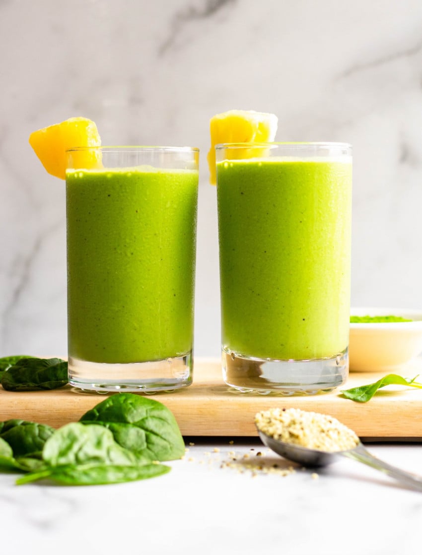 Two pineapple matcha green smoothies in glasses on wooden cutting board with ingredients surrounding the cups