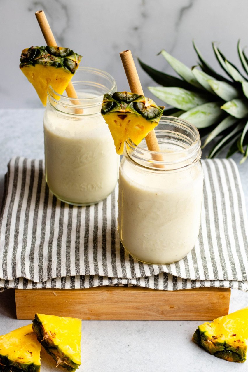 Two pina colada smoothies in mason jars with bamboo straws and fresh pineapple garnish