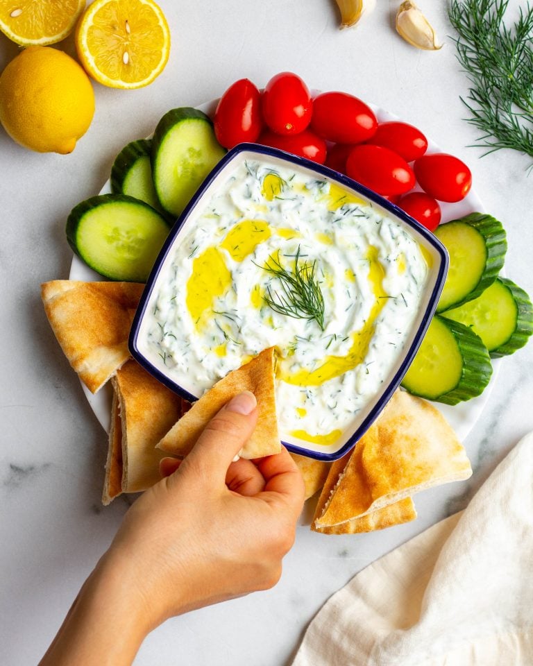 Hand dipping a piece of pita bread into homemade tzatziki sauce surrounded by a small plate of veggies