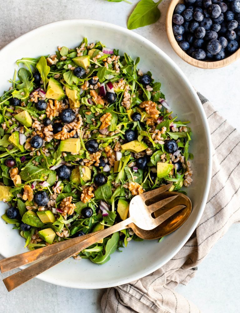 Large bowl of blueberry basil salad with gold serving spoons and a bowl of blueberries on the side