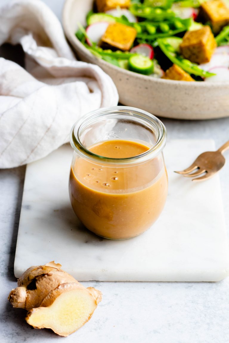 Jar of sesame ginger miso dressing on a small marble board with a gold fork and salad in the background
