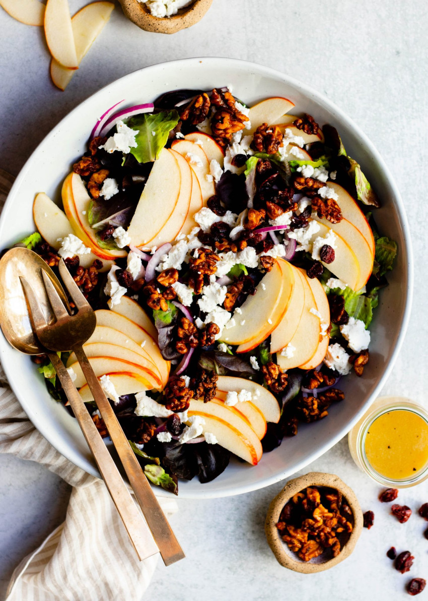 Apple cranberry walnut salad in large salad bowl with gold mixing spoons, surrounded by small bowls of ingredients and salad dressing
