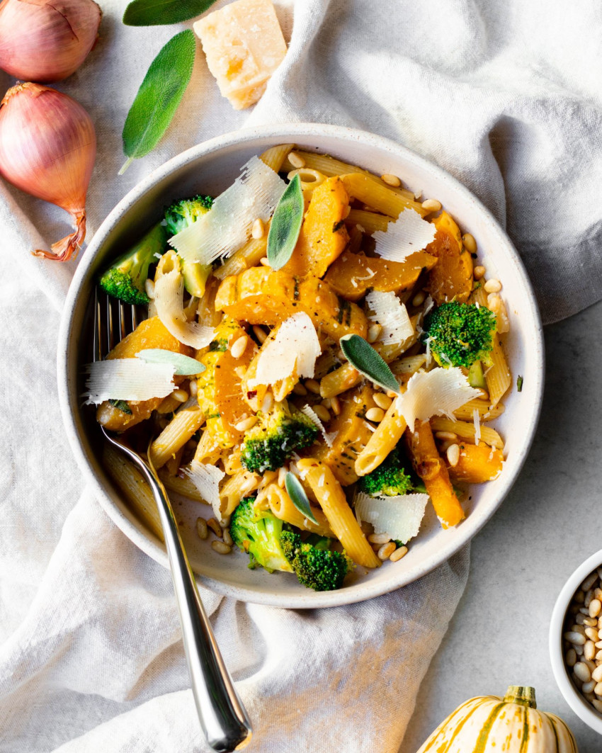 Bowl of delicata squash pasta with a silver fork on a linen napkin, surrounded by different ingredients