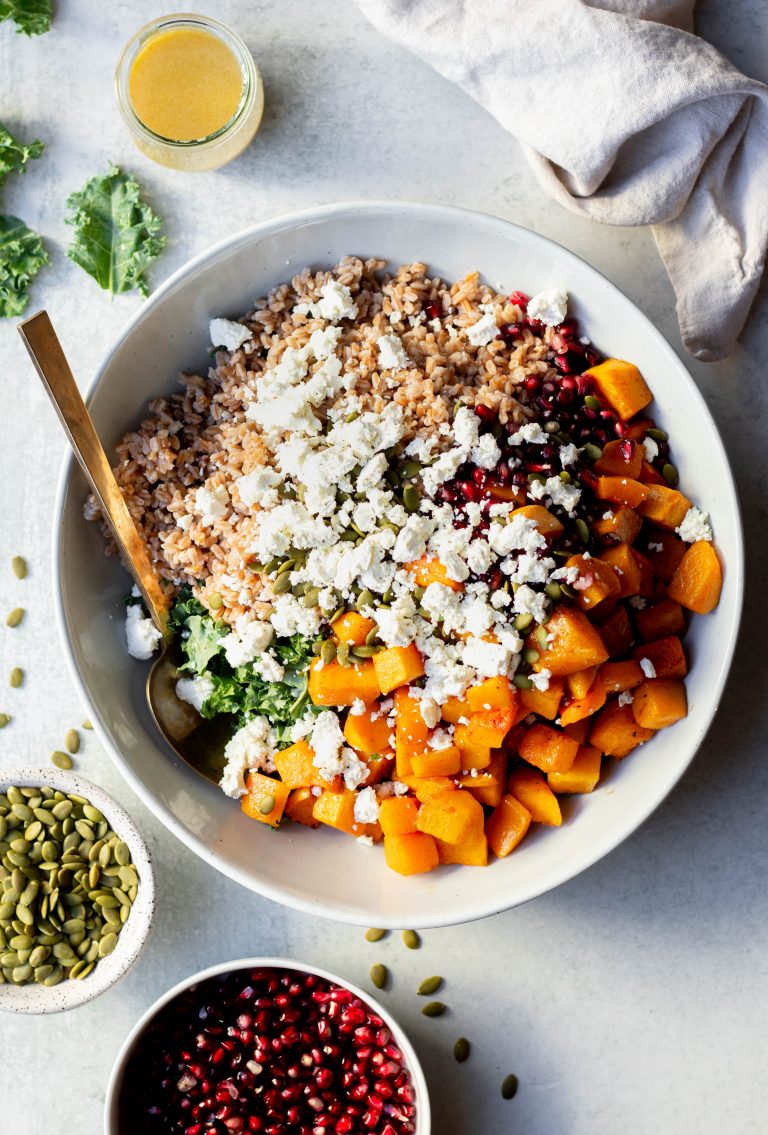 Large bowl of fall farro salad with gold mixing spoon surrounded by bowls of ingredients