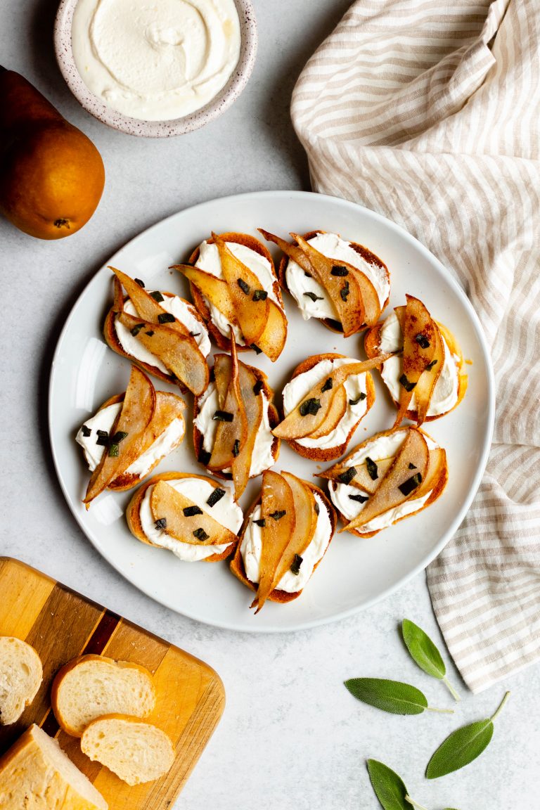 Plate of pear crostini with sage leaves scattered on grey backdrop beside a loaf of sliced French bread