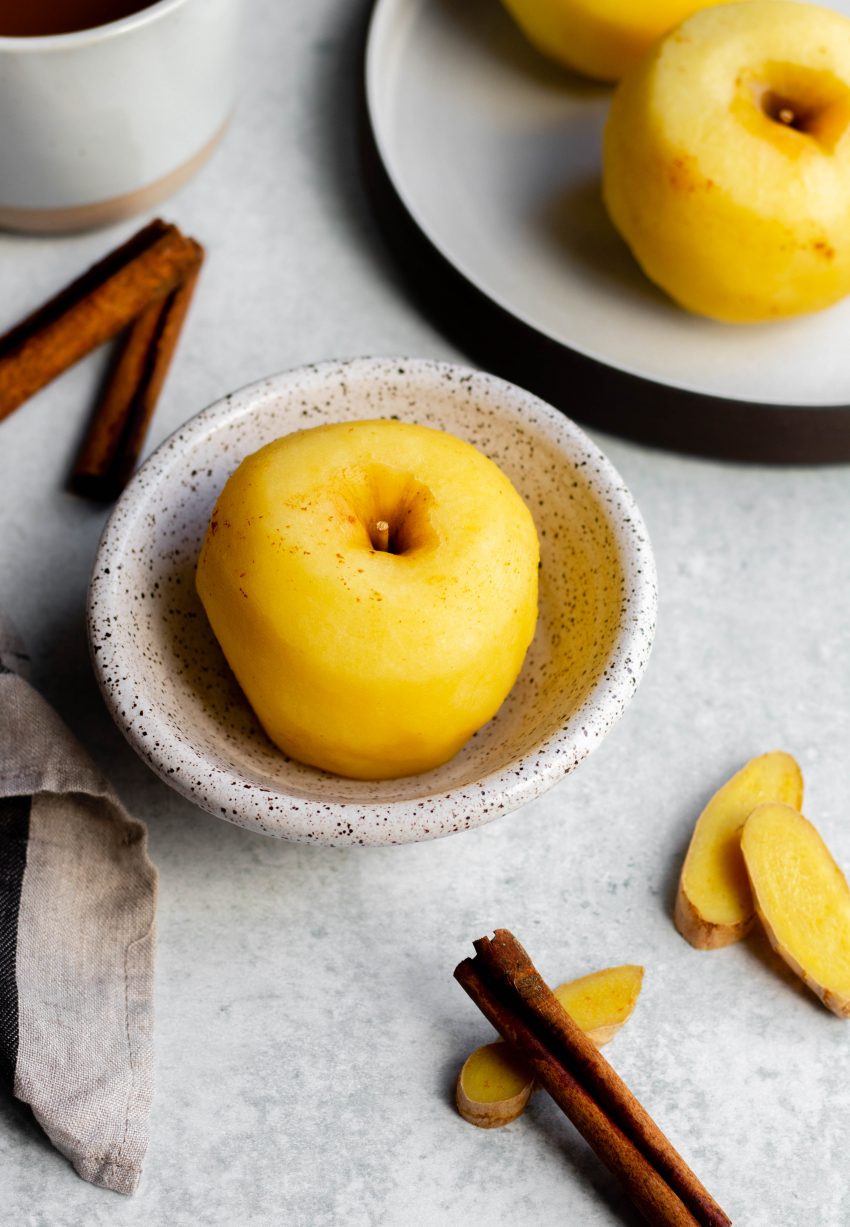 Poached apple in a small bowl, next to a plate of other poached apples, with cinnamon sticks and ginger slices surrounding