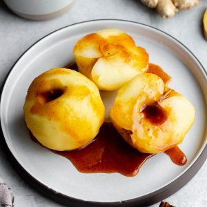 Poached apples on a grey plate drizzled with syrup, cinnamon sticks and fresh ginger surrounding the plate