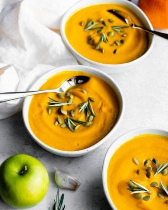 Three bowls of pumpkin apple soup garnished with pepitas and rosemary