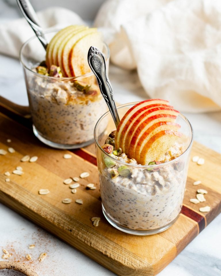 Two glasses filled with chai latte overnight oats topped with apple slices on a wooden cutting board