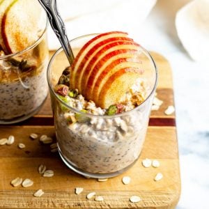 close up of a glass of chai latte overnight oats with fresh apple slices and a silver spoon on wood cutting board