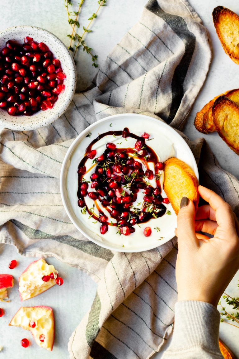 Bowl of honey whipped goat cheese with pomegranate on grey towel with crostini surrounding the bowl