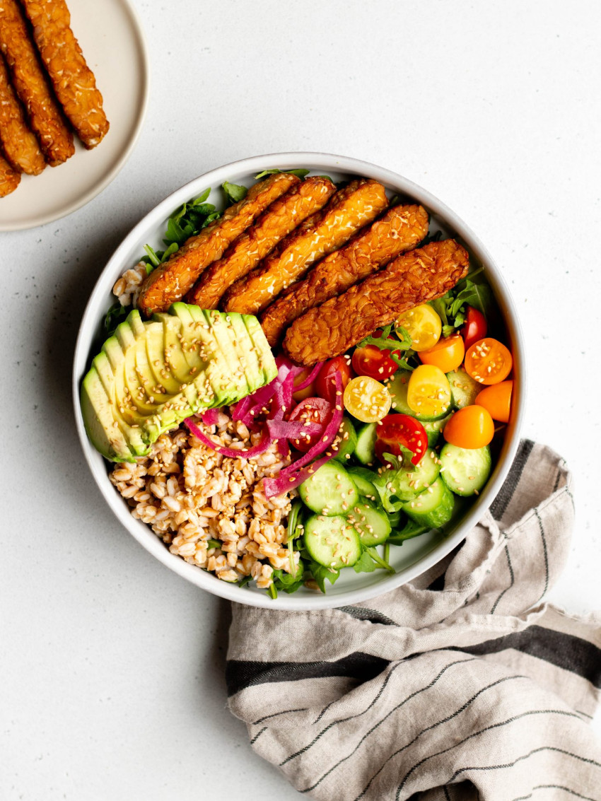 Colorful grain bowl with air fried tempeh strips with grey towel on a white backdrop