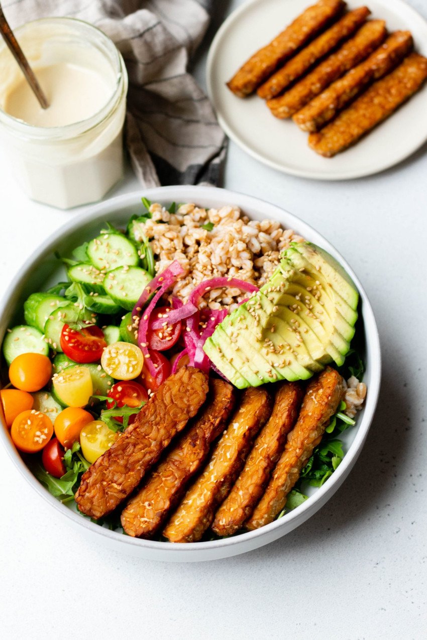 Colorful grain bowl with air fried tempeh strips on a white backdrop