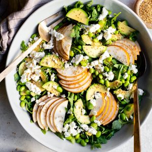 Large bowl of asian pear salad with gold serving spoons, with an asian pear, small bowl of sesame seeds, and small jar of dressing surrounding
