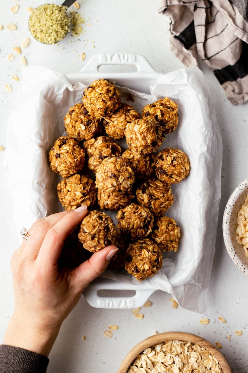 Hand grabbing a no bake granola ball from a white dish, surrounded by some bowls of ingredients