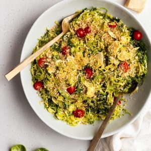Big bowl of shredded Brussels sprouts salad with gold serving spoons surrounded by ingredients on a grey backdrop