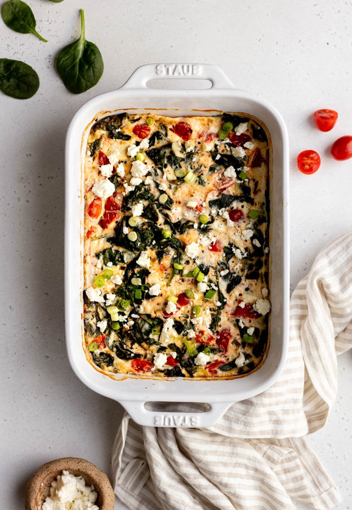 Baked veggie egg white frittata in a white ceramic dish on a grey backdrop with a linen dish cloth and some veggie ingredients surrounding the dish