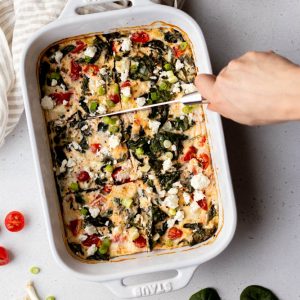 Hand cutting veggie egg white frittata into squares in a white ceramic baking dish on a grey backdrop