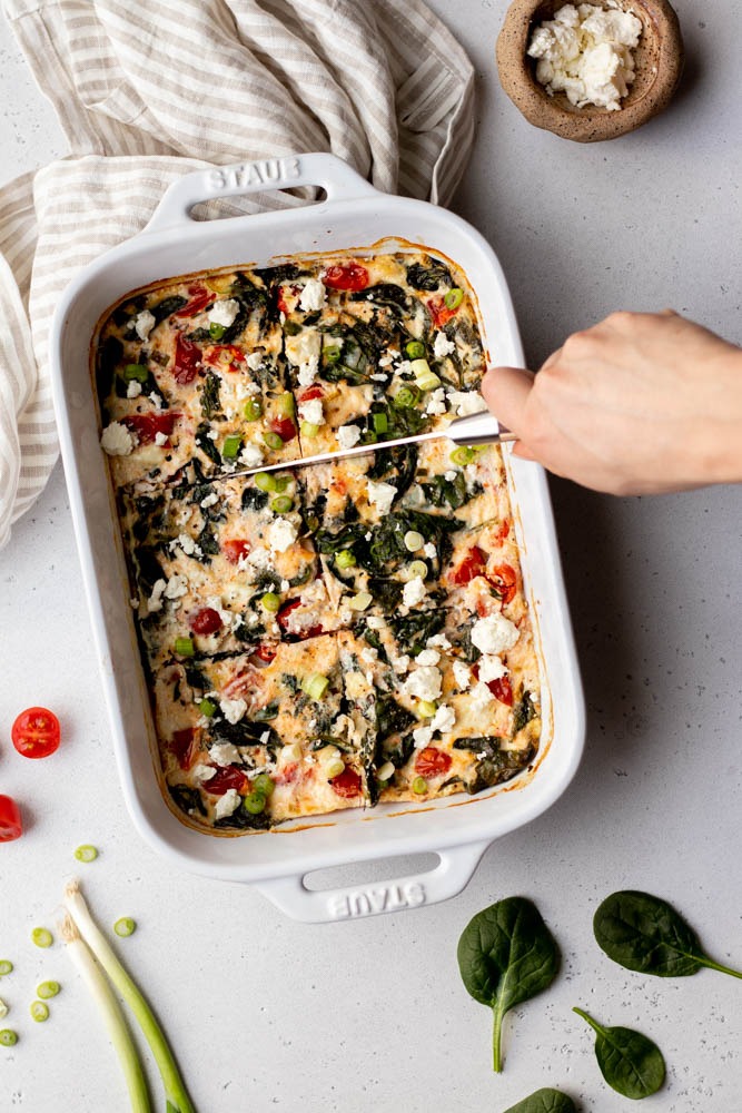 Hand cutting veggie egg white frittata into squares in a white ceramic baking dish on a grey backdrop