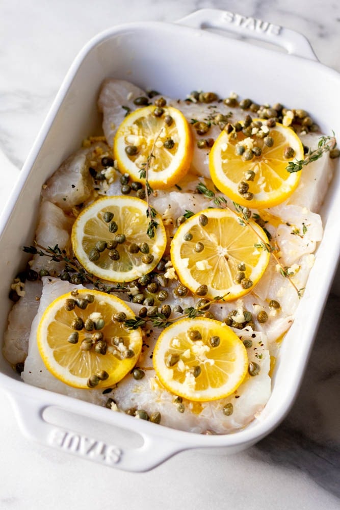 Close up of cod topped with lemon slices, capers, and herbs in a white baking dish