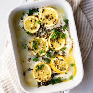Baked cod fish topped with lemon slices, capers, and herbs in a white baking dish