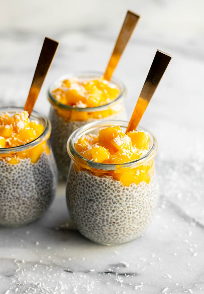 three cups of mango coconut chia pudding with gold spoons on a white marble backdrop
