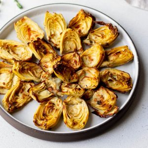 Plate with air fried artichoke hearts on a grey backdrop, surrounded by a wedge of parmesan cheese and sprigs of thyme