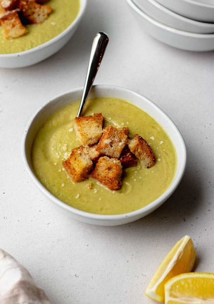 close up of a bowl of broccoli leek soup with a silver spoon. Soup is topped with croutons and there is sliced lemon in the corner.