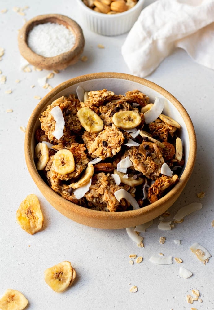 Close up of a bowl of granola topped with banana chips, coconut shreds, and ingredients scattered around a grey backdrop