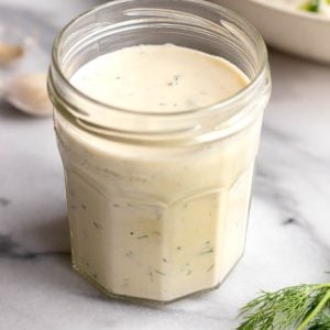 Glass jar of Greek yogurt salad dressing on a white marble tabletop with a sprig of fresh dill