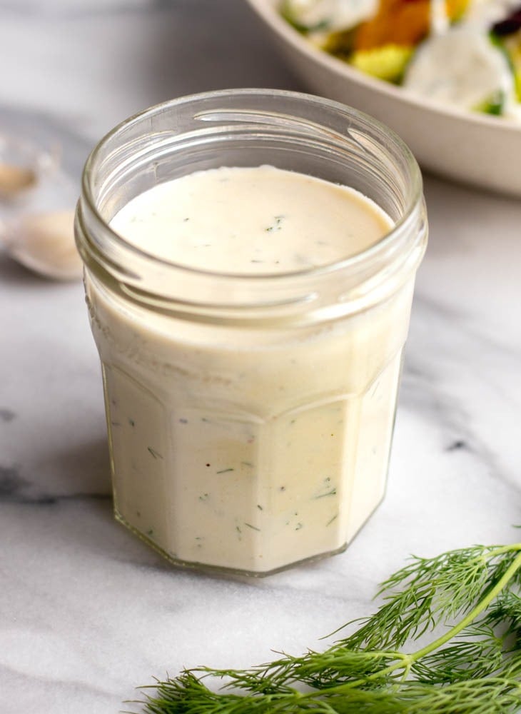 Glass jar of Greek yogurt salad dressing on a white marble tabletop with a sprig of fresh dill