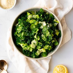 Kale and romaine salad in a bowl sitting on a white dish towel, surrounded by some of the salad ingredients and gold salad spoons