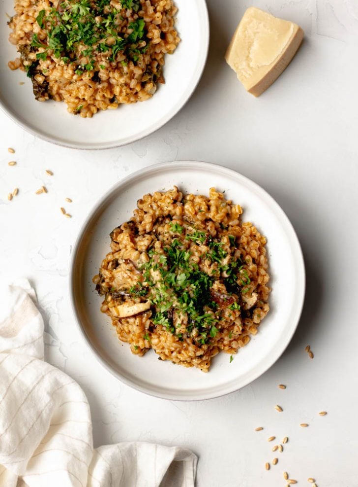 Two bowls of farro mushroom risotto with a hunk of parmesan cheese on the side, a neutral dish towel, on a light grey backdrop