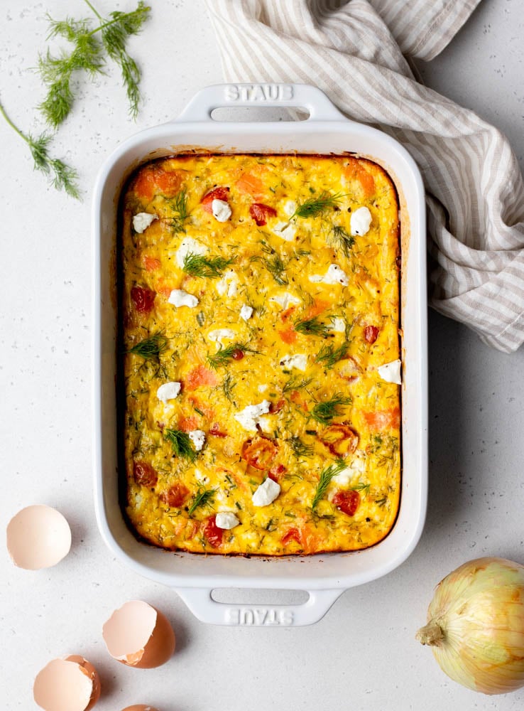 Smoked salmon frittata in a white baking dish with raw ingredients surrounding the dish on a light grey backdrop