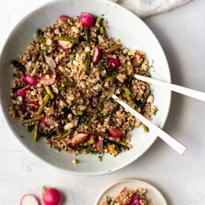Large bowl of spring farro salad with gold mixing spoons with a small plate of salad on a white backdrop
