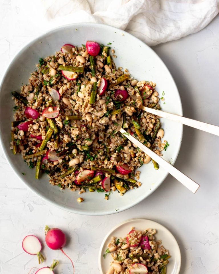 Large bowl of spring farro salad with gold mixing spoons with a small plate of salad on a white backdrop